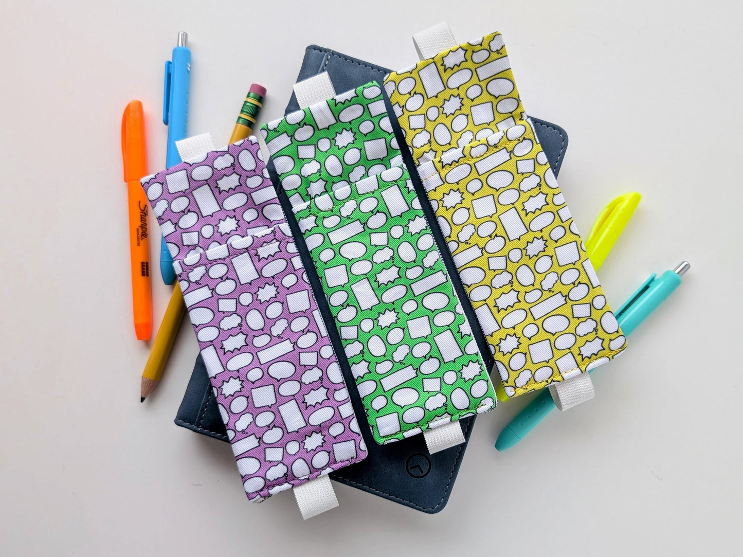 Three patterned bookmarks on top of a notebook with pens and pencils on a light gray background